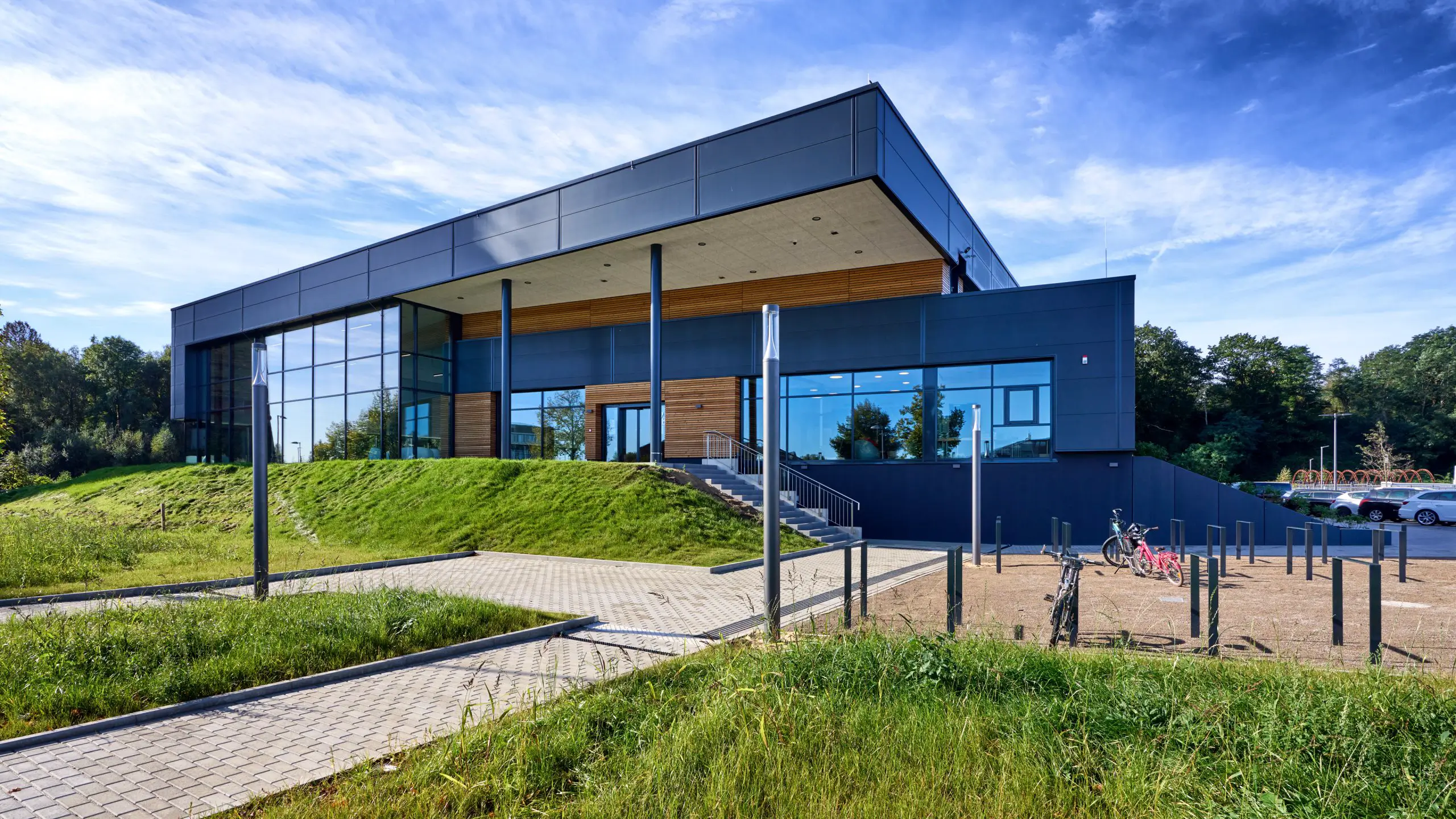 Moderne, zweigeschossige Gebäudefront mit großen Glasflächen und schwarzer Fassade am Standort Annabad in Alsdorf. Vor dem Gebäude verlaufen Wege, Rasenflächen und ein Fahrradstellplatz. Der Himmel ist blau mit leichten Wolken.